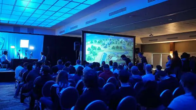 A packed conference audience watching a large stage screen