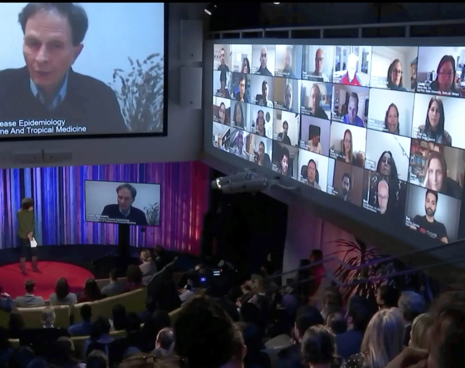 Hybrid conference with a remote speaker and attendee grid on the stage screen above a live audience