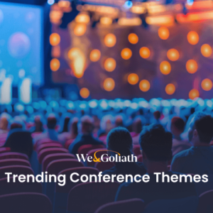 Ignite Passion with These Trending Conference Themes Audience at a conference with focus on the stage, lights dimmed, text overlay "trending conference themes" and logo "we&goliath.