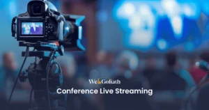 Best Practices for Live Streaming Your Conference A video camera is set up to live stream a conference, with a blurred audience and presenter in the background, and the text "We & Goliath Conference Live Streaming" displayed at the bottom.