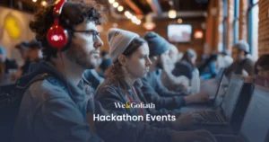 Hackathon Guide: Tech Events You Can't Miss People working on laptops at a hackathon event in a spacious indoor venue; one individual is wearing red headphones. Text on image: "We & Goliath Hackathon Events.