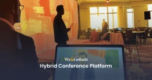 Hybrid Conference Tools: 2024's Best Platforms People are attending a conference; one speaker stands on stage holding a paper, another person sits in front of a laptop displaying the We & Goliath logo and "Hybrid Conference Platform" text.
