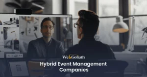 The Hybrid Event Management Company That Delivers Results A person sits at a desk, engaging in a video call with another individual shown on a computer screen. Text overlay reads "We & Goliath Hybrid Event Management Companies.