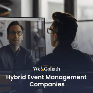 A person sits at a desk, engaging in a video call with another individual shown on a computer screen. Text overlay reads "We & Goliath Hybrid Event Management Companies.