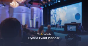 Crafting a Standout Hybrid Event: A Guide for Event Planners Audience members facing a stage with two people seated in discussion, a large screen behind them displaying visuals. A video camera on the right side is recording the event. Text overlay reads, "We & Goliath Hybrid Event Planner.