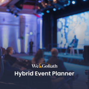 Audience members facing a stage with two people seated in discussion, a large screen behind them displaying visuals. A video camera on the right side is recording the event. Text overlay reads, "We & Goliath Hybrid Event Planner.