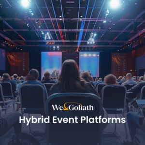 A large audience seated in a conference hall, facing a stage with a presentation screen. Text on the image reads "We & Goliath Hybrid Event Platforms.