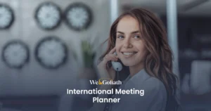 International Meeting Planner Tool for Scheduling Across Time Zones A person smiling while talking on a phone with "We & Goliath International Meeting Planner" text overlaid on the image. Rows of clocks are visible on the wall in the background.