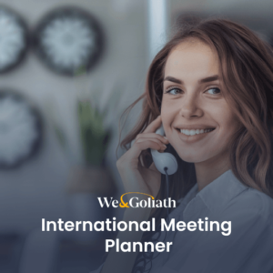International Meeting Planner Tool for Scheduling Across Time Zones A person smiling while talking on a phone with "We & Goliath International Meeting Planner" text overlaid on the image. Rows of clocks are visible on the wall in the background.