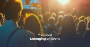 Effective Event Planning and Attendee Management A large audience is seated facing a stage set for a presentation. The image is lit warmly from the sunset or bright lights. Text overlay reads: "We & Goliath: Managing an Event".