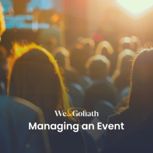 Effective Event Planning and Attendee Management A large audience is seated facing a stage set for a presentation. The image is lit warmly from the sunset or bright lights. Text overlay reads: "We & Goliath: Managing an Event".