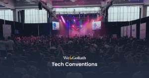 Discover the Best Tech Conferences and Events in 2024 A large audience attends a tech convention in a spacious modern venue with stage lighting and multiple screens. The text "We & Goliath Tech Conventions" is displayed at the bottom.