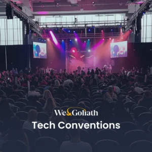 Discover the Best Tech Conferences and Events in 2024 A large audience attends a tech convention in a spacious modern venue with stage lighting and multiple screens. The text "We & Goliath Tech Conventions" is displayed at the bottom.