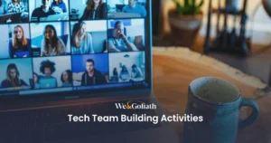 Tech Team Building Activities: Uniting Virtual Teams at Work A laptop screen shows a video call with multiple participants. Text on the image reads, "We & Goliath Tech Team Building Activities." A mug sits on the table in front of the laptop.