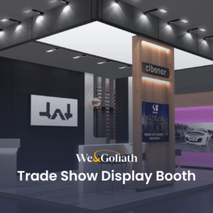 A trade show display booth featuring the We & Goliath logo, with various branded sections, promotional materials, and product displays under bright overhead lighting.