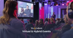 Virtual Events vs. Hybrid Events: What's the Difference? An audience sits in a conference room wearing headphones and watching presenters on a stage with a large screen. The text "We & Goliath Virtual & Hybrid Events" is overlaid on the image.