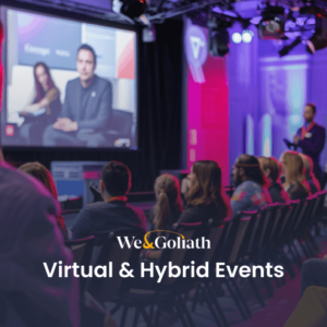 Virtual Events vs. Hybrid Events: What's the Difference? An audience sits in a conference room wearing headphones and watching presenters on a stage with a large screen. The text "We & Goliath Virtual & Hybrid Events" is overlaid on the image.