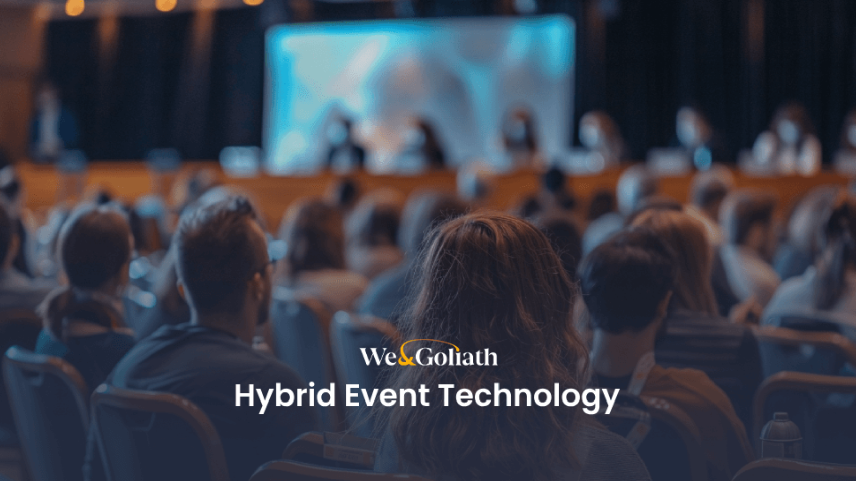 An audience watches a presentation on a stage with the text "We & Goliath Hybrid Event Technology" overlayed at the bottom.