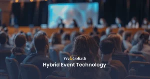 Understanding Hybrid Events: Technology and Best Practices An audience watches a presentation on a stage with the text "We & Goliath Hybrid Event Technology" overlayed at the bottom.