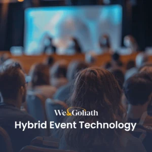 Understanding Hybrid Events: Technology and Best Practices An audience watches a presentation on a stage with the text "We & Goliath Hybrid Event Technology" overlayed at the bottom.