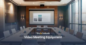 Meeting Room Tech: Video Conference Equipment Guide A modern conference room equipped with a large screen displaying a presentation, surrounded by tables and chairs set up for a video meeting. Text reads "We & Goliath Video Meeting Equipment.