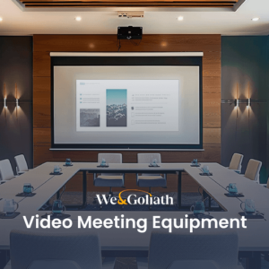 A modern conference room equipped with a large screen displaying a presentation, surrounded by tables and chairs set up for a video meeting. Text reads "We & Goliath Video Meeting Equipment.