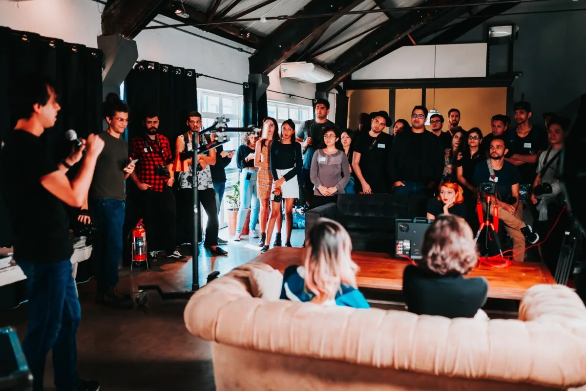 Speaker with a microphone addressing a standing audience in a loft venue while a camera crew films the session