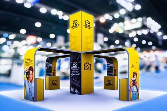 Large yellow and black four-sided island trade show booth for a camera rental brand on a convention hall floor