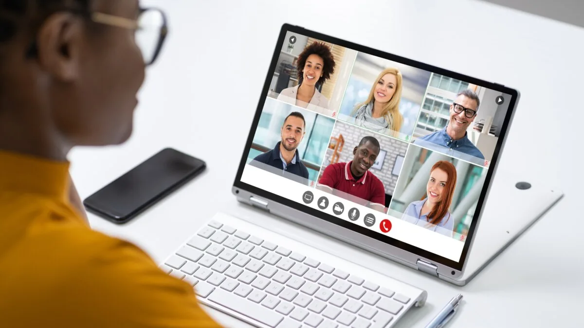 Person in a yellow top joining a six-person video call on a laptop, seen over their shoulder